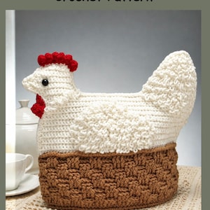 May include: A crochet tea cozy shaped like a white hen with a red comb and wattle, sitting on a brown basket-weave base. The text Nesting Hen Tea Cozy Crochet Pattern is displayed at the top of the image in a clear, dark font.