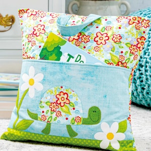May include: A decorative pillow with a light blue pocket featuring a turtle applique with floral details. The pillow has a floral pattern on the top and a green border. A book is in the pocket.