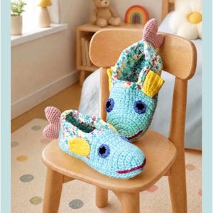 Peut inclure: Une paire de chaussons en crochet bleu clair en forme de poisson, avec des nageoires latérales jaunes, des nageoires caudales roses, des yeux circulaires bleu foncé et une bouche rose foncé. L'intérieur est doublé d'un tissu floral. Texte Tini Dancer Vintage Collection.