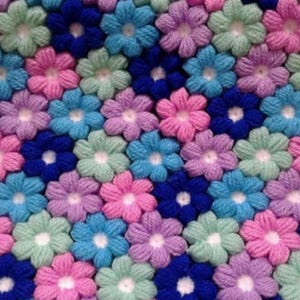 May include: Close-up of a handmade textile featuring a repeating pattern of crocheted flowers. The flowers are in shades of pink, blue, green, and purple, with a dark blue background. The texture is soft and inviting, suggesting a cozy blanket or decorative item.