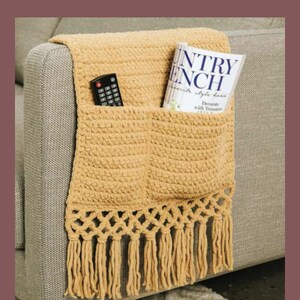 May include: A mustard yellow crocheted couch armrest organizer with two pockets and a fringe trim. A remote control and a book titled "Country French" are in the pockets. The organizer is draped over a gray couch arm.