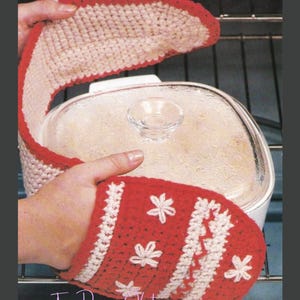 May include: A double-sided crochet oven mitt featuring a red and cream color scheme with embroidered white floral accents. The mitt has a long, connected design with red trim and is positioned over a square glass casserole dish with a clear lid.
