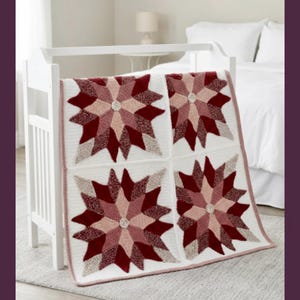 May include: A square crochet blanket featuring four large star motifs in shades of deep red, burgundy, and soft pink on a white background. Each star has a small, light-colored circular center. The blanket has a simple white border with a thin pink edge.