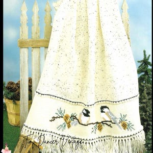 May include: A cream-colored crocheted throw blanket featuring a speckled texture and a decorative border. The border displays a cross-stitched pattern of two chickadees perched on pine branches with pinecones, finished with long fringe.