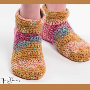 May include: A pair of ankle-length crochet slippers featuring a variegated yarn pattern in shades of mustard yellow, magenta, teal, and cream. The slippers have a thick, textured knit construction with a folded cuff at the top and the text Tini Dancer.