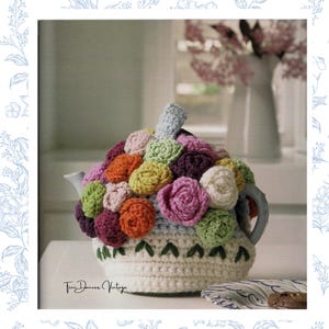 May include: A cream-colored crochet tea cozy featuring a cluster of multi-colored yarn flowers in shades of pink, orange, purple, green, and white. The base includes a row of green embroidered leaf accents and the text Tini Dancer Vintage.