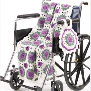 Op de afbeelding: Een gehaakte deken en een bijpassende ronde tas met een bloemmotief in wit, saliegroen en paars garen. De deken is gedrapeerd over de zitting en rugleuning van een metalen rolstoel, terwijl de ronde tas aan de armleuning hangt.