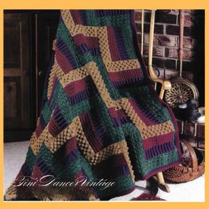 May include: A crocheted blanket featuring a geometric pattern of interlocking rectangular blocks in deep forest green, burgundy, and tan. The texture consists of alternating smooth and raised bobble stitch sections throughout the design.