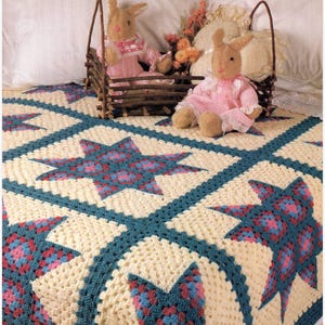 Könnte beinhalten: Eine cremefarbene Häkeldecke mit einem wiederkehrenden Muster aus großen achtstrahligen Sternen in Blaugrün, Rosa und Blau. Zwei plüschige, hellbraune Hasenpuppen in rosa-weißer Kleidung sitzen in einem rustikalen Korb aus Zweigen auf der Decke.