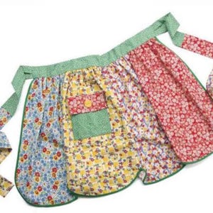 Könnte beinhalten: Eine bunte Halbschürze mit drei Abschnitten, die jeweils ein anderes Blumenmuster aufweisen. Die Schürze hat einen grünen Pünktchenbund und Bindebänder sowie eine grüne Tasche. Die Muster umfassen blaue, gelbe und rote Blumen.