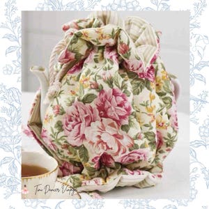 Könnte beinhalten: Eine gesteppte Teewärmer-Hülle aus Stoff mit cremefarbenem Hintergrund und einem Blumenmuster aus rosa Rosen, grünen Blättern und gelben Akzenten. Die Hülle hat einen Kordelzug mit dicken weißen Seilbändern und ein gestreiftes Innenfutter.