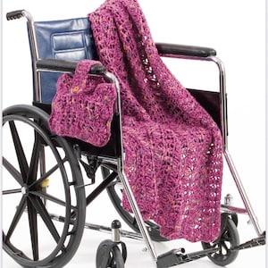 May include: A magenta crochet blanket with a variegated yarn texture draped over a wheelchair, accompanied by a matching small rectangular crochet pouch with a button closure attached to the armrest of the wheelchair.
