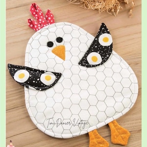 May include: A fabric potholder shaped like a chicken with a white body featuring a black honeycomb pattern. It has a red and white polka dot comb, black button eyes, an orange beak, black wings with yellow egg prints, and orange feet.