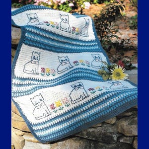 May include: A handmade crochet blanket featuring a repeating pattern of cartoon cats and flowers. The blanket has alternating stripes of white and blue, with a dark blue border. The cat and flower designs are embroidered in black and various colors.