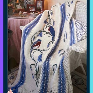 May include: A cream-colored crocheted throw blanket featuring a central design of blue and red birds perched on floral branches, bordered by vertical blue and white textured stripes, and finished with long fringe along the bottom edge.