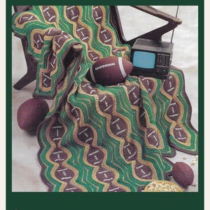 May include: A crochet blanket featuring a repeating pattern of brown football shapes with white laces, separated by wavy green and tan stripes. The blanket is draped over a chair alongside three brown footballs and a small vintage television set.