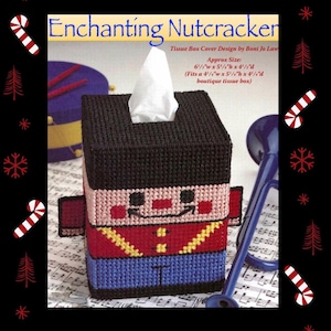 May include: A square plastic canvas tissue box cover shaped like a nutcracker with a black hat, pink face, red jacket with yellow trim, and blue pants. The text reads Enchanting Nutcracker, Tissue Box Cover Design by Boni Jo Law, Approx Size 6 1/2"w x 5 1/4"h x 4 1/2"d.