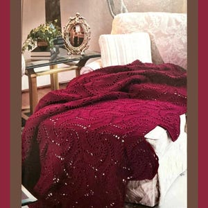 May include: A deep burgundy knitted throw blanket featuring an intricate, repeating swirl pattern with openwork details. The textile is draped over a light-colored upholstered chair, showcasing its textured surface and rich, dark red color.