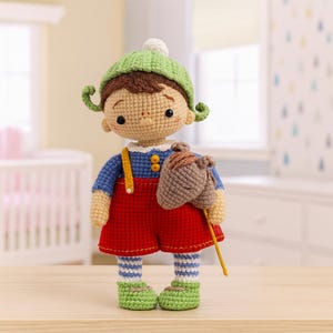 Puede incluir: Mu&ntilde;eca de ganchillo hecha a mano con gorro verde, camisa azul, pantalones cortos rojos y zapatos verdes. La mu&ntilde;eca tiene un juguete en forma de bellota marr&oacute;n y est&aacute; sobre una superficie de madera. La mu&ntilde;eca tiene pelo rizado verde y un pomp&oacute;n blanco en el gorro.