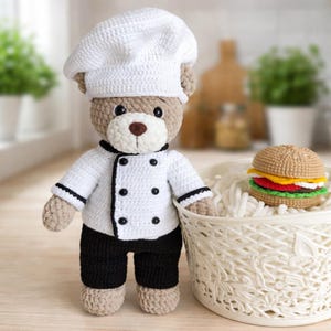 K&ouml;nnte beinhalten: Ein geh&auml;kelter Teddyb&auml;r, der als Koch gekleidet ist und einen wei&szlig;en Hut und eine Jacke mit schwarzen Verzierungen und Kn&ouml;pfen sowie eine schwarze Hose tr&auml;gt. Ein geh&auml;kelter Hamburger liegt in einem wei&szlig;en Korb. Der B&auml;r ist hellbraun mit einer braunen Nase.