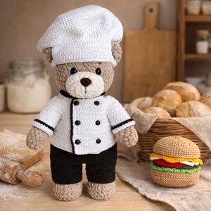 K&ouml;nnte beinhalten: Ein geh&auml;kelter Teddyb&auml;r in Kochkleidung, mit wei&szlig;er Kochm&uuml;tze und Jacke mit schwarzen Details und schwarzer Hose. Ein geh&auml;kelter Burger liegt daneben. Im Hintergrund ein Korb mit Brot.