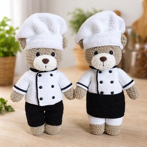 K&ouml;nnte beinhalten: Zwei geh&auml;kelte Teddyb&auml;ren als K&ouml;che verkleidet. Jeder B&auml;r tr&auml;gt eine wei&szlig;e Kochm&uuml;tze, eine wei&szlig;e Jacke mit schwarzen Kn&ouml;pfen und Besatz sowie eine schwarze Hose oder eine schwarze Sch&uuml;rze. Die B&auml;ren halten sich an den H&auml;nden, mit braunen Augen und Nasen.