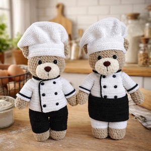 Könnte beinhalten: Zwei gehäkelte Teddybären als Köche verkleidet. Jeder Bär trägt eine weiße Kochmütze, eine weiße Kochjacke mit schwarzen Verzierungen und Knöpfen sowie eine schwarze Hose oder eine schwarze Schürze. Die Bären sind hellbraun mit braunen Nasen und schwarzen Augen.