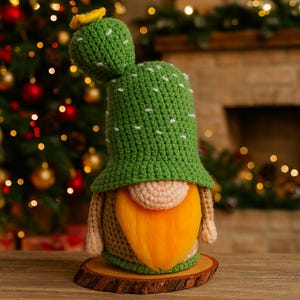 Puede incluir: Figura de gnomo cactus de ganchillo hecha a mano. El gnomo lleva un gorro verde con detalles blancos y una flor amarilla. Tiene una cara beige y una barba naranja. El gnomo se exhibe sobre una rodaja de madera.