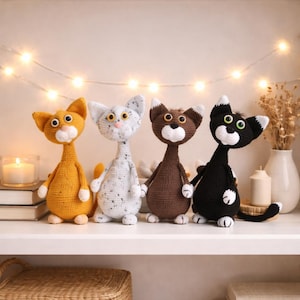 Könnte beinhalten: Vier handgefertigte Häkelkatzenpuppen in Orange, Weiß mit grauen Sprenkeln, Braun und Schwarz. Jede Katze hat große Augen, eine weiße Schnauze und einen langen Hals. Sie stehen auf einem weißen Regal.