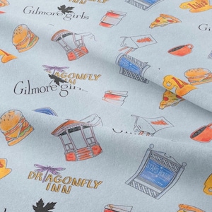Pu&ograve; includere: Tessuto azzurro con un motivo ripetuto di cibo, edifici e testo. Il testo include "Gilmore Girls", "Dragonfly Inn" e "Stars Hollow". Le illustrazioni sono in arancione, rosso e giallo.