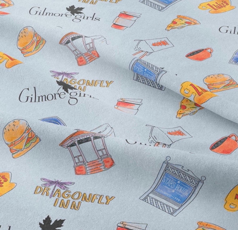 Pu&ograve; includere: Tessuto azzurro con un motivo ripetuto di cibo, edifici e testo. Il testo include "Gilmore Girls", "Dragonfly Inn" e "Stars Hollow". Le illustrazioni sono in arancione, rosso e giallo.