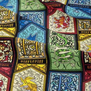 May include: Fabric with a repeating pattern of stained-glass style crests. Each crest features a house name: Gryffindor, Hufflepuff, Ravenclaw, and Slytherin. The crests are in various colors, including red, gold, green, and blue, with decorative borders and lettering.