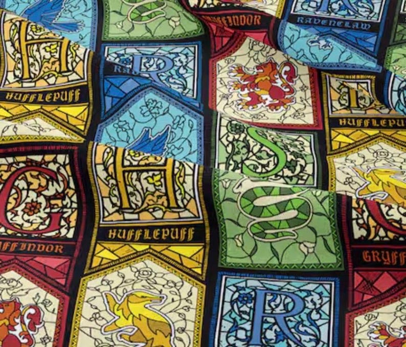 May include: Fabric with a repeating pattern of stained-glass style crests. Each crest features a house name: Gryffindor, Hufflepuff, Ravenclaw, and Slytherin. The crests are in various colors, including red, gold, green, and blue, with decorative borders and lettering.
