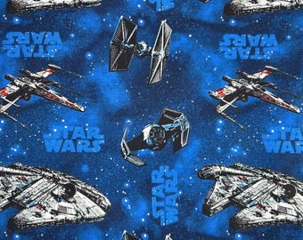 May include: Blue fabric featuring a Star Wars theme. The design includes illustrations of the Millennium Falcon, X-wing fighters, and TIE fighters, along with the Star Wars logo. The background is a dark blue with white star-like details.