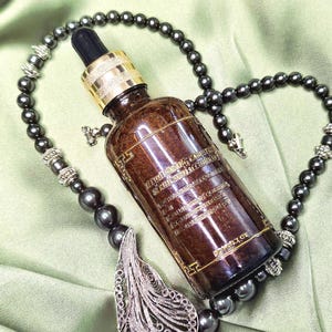 May include: A brown glass bottle with a dropper, gold cap, and text, resting on a green surface. The bottle is surrounded by a dark gray beaded necklace with a silver filigree pendant.