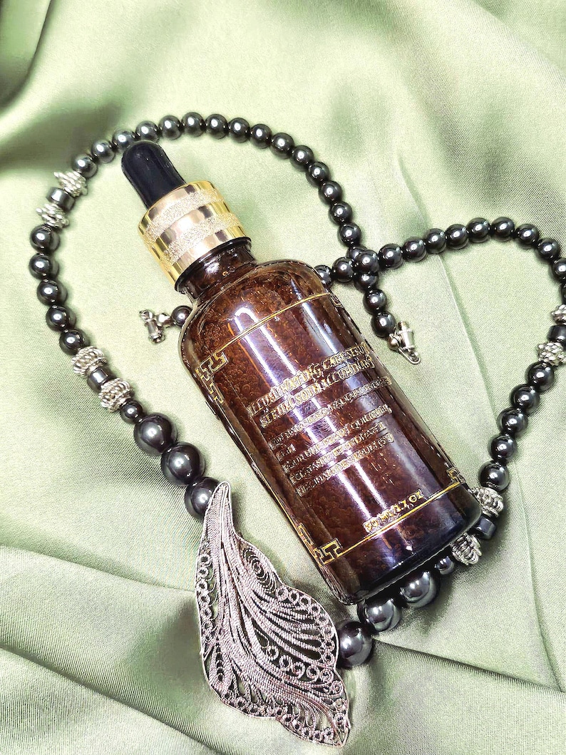 May include: A brown glass bottle with a dropper, gold cap, and text, resting on a green surface. The bottle is surrounded by a dark gray beaded necklace with a silver filigree pendant.