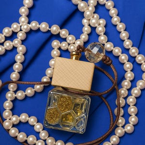 May include: A small, clear glass perfume bottle with a wooden cap and gold glitter inside, on blue fabric. A brown cord is attached to the bottle, and a strand of white pearls surrounds the bottle.