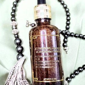 May include: A brown glass bottle with a gold-colored cap and dropper, featuring text on the label. The bottle is surrounded by a dark gray beaded necklace and a decorative pendant. The bottle appears to contain a serum.