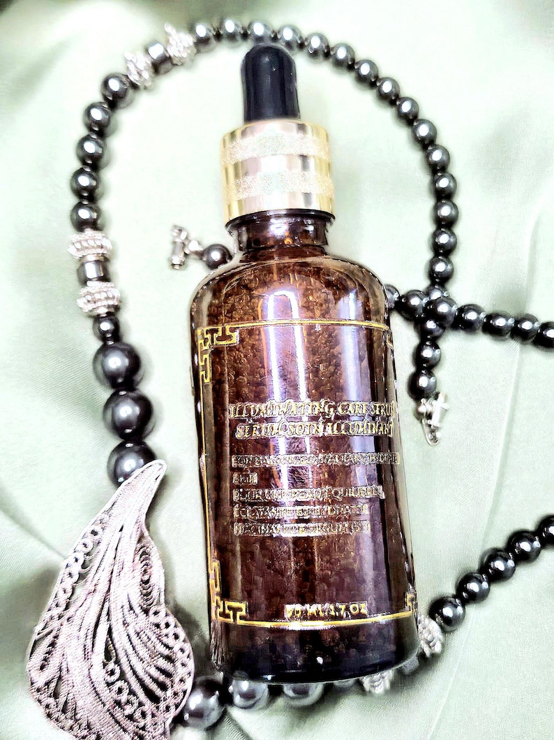 May include: A brown glass bottle with a gold-colored cap and dropper, featuring text on the label. The bottle is surrounded by a dark gray beaded necklace and a decorative pendant. The bottle appears to contain a serum.