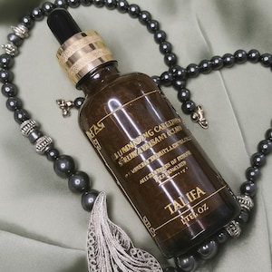 May include: A brown glass bottle with a gold cap and black dropper, labeled "TALIFA". The bottle is surrounded by a dark gray beaded necklace with a silver feather pendant. The background is a light green fabric.