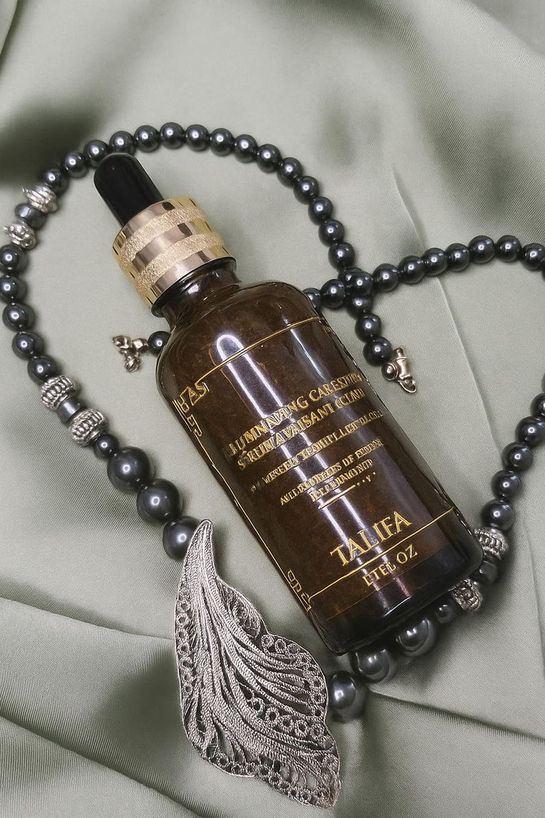 May include: A brown glass bottle with a gold cap and black dropper, labeled "TALIFA". The bottle is surrounded by a dark gray beaded necklace with a silver feather pendant. The background is a light green fabric.
