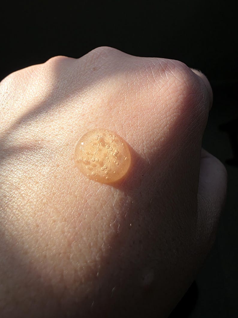 May include: A close-up shot of a hand with a small, round, amber-colored droplet of liquid on the skin. The droplet has a slightly bubbly texture and reflects the light. The skin is light-toned and the background is dark.