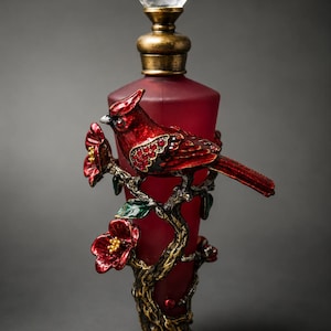 May include: A frosted deep red glass perfume bottle featuring a decorative metal overlay of a red cardinal bird perched on a branch with red flowers. The bottle is topped with a clear faceted crystal stopper and a metallic gold-toned neck.