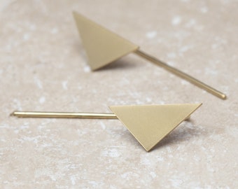 May include: Two gold-colored earrings. Each earring features a triangle shape attached to a thin, straight post. The earrings are displayed on a light-colored, textured surface.