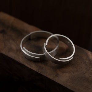 Peut inclure: Deux bagues réglables en argent sont présentées sur une surface en bois foncé. Les bagues ont un design moderne et minimaliste avec un espace dans le bracelet pour l'ajustement de la taille. Les bagues sont en métal brossé.