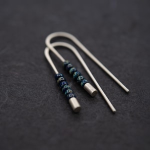 Op de afbeelding: Zilverkleurige oorbellen met een uniek U-vormig ontwerp. Elke oorbel heeft een reeks donkerblauwe en turquoise kralen aan de basis. De donkere achtergrond benadrukt het ontwerp en de kleuren.