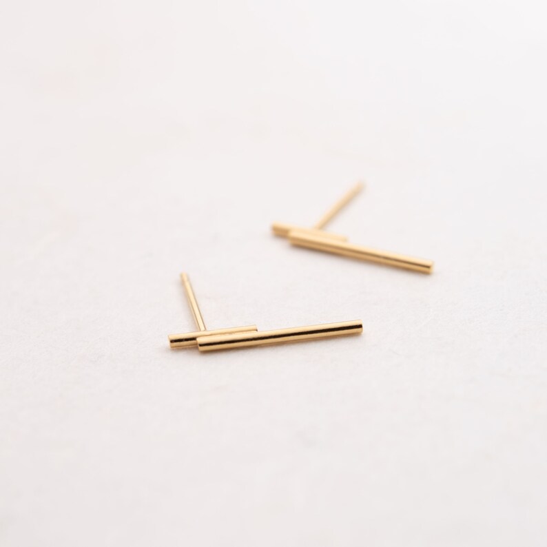May include: A pair of gold bar stud earrings. The earrings are made of two thin gold bars that are stacked on top of each other.