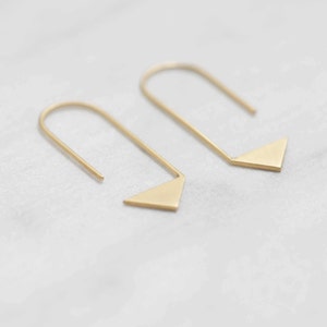 Puede incluir: Un par de pendientes de triángulo de oro con un diseño simple y minimalista. Los pendientes están hechos de un alambre delgado que forma un bucle y luego se extiende en una pequeña forma triangular.