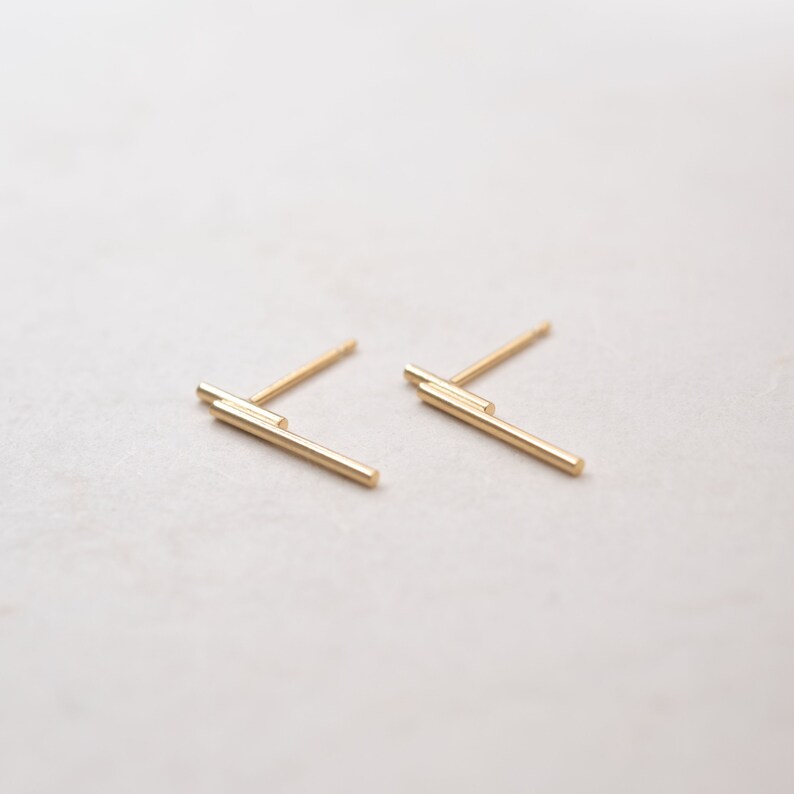 May include: A pair of gold bar stud earrings. The earrings are made of two thin gold bars that are stacked on top of each other.