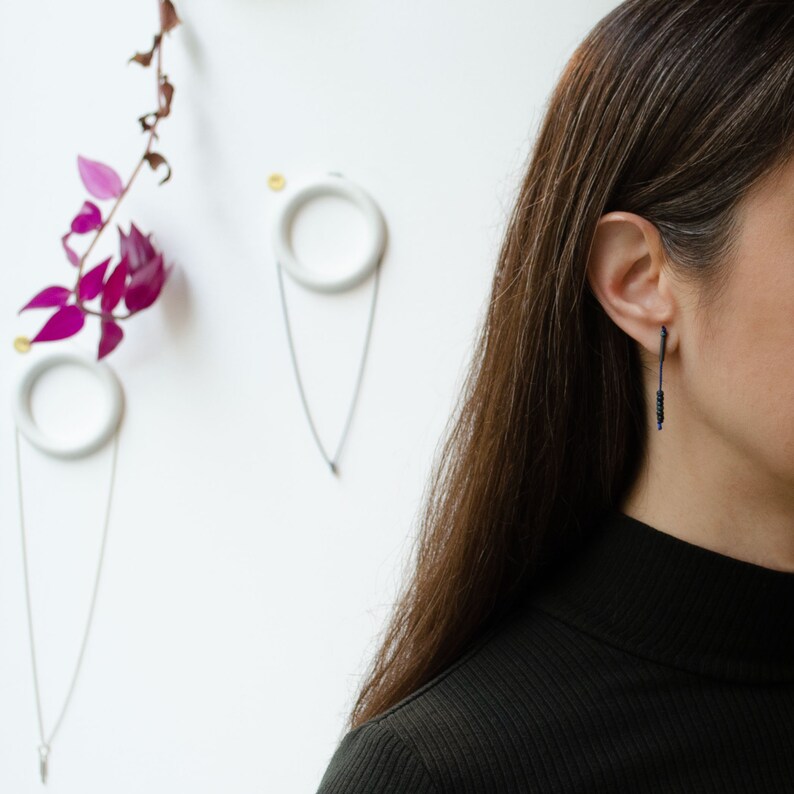 Woman half face wearing a pendant earring of 38 mm length handmade by A g J c from an oxidized sterling silver tube with a blue silk cord threaded through the center for hanging Miyuki beads in Picasso cobalt blue.